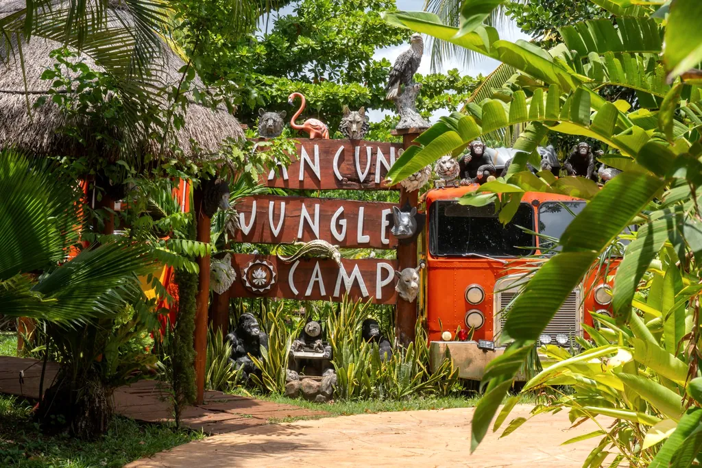 Letrero de madera de la entrada de Cancún Jungle Camp entre hojas de plátano, con el camión naranja detrás